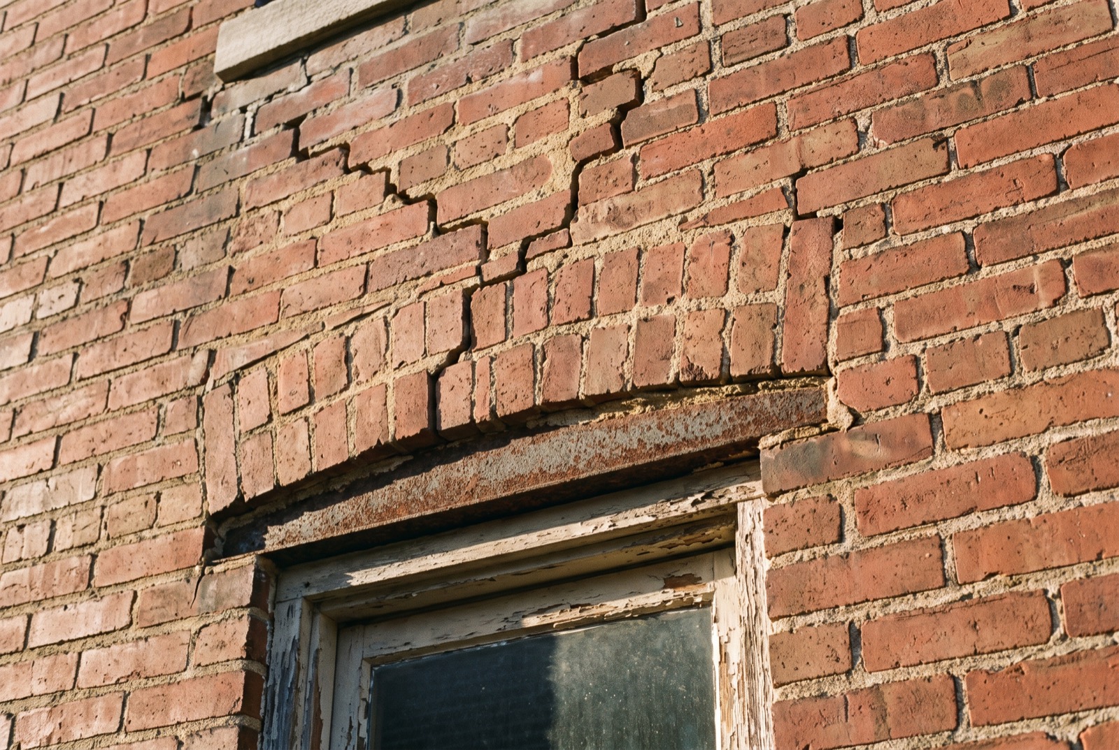 A stair-step crack in a brick wall above a rusted window lintel